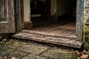 Close-up photograph of a weathered wooden door threshold with peeling paint, stone pavers in front